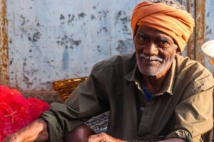 A senior Indian man wearing an orange turban smiles warmly while seated outdoors in Cumbum, India.